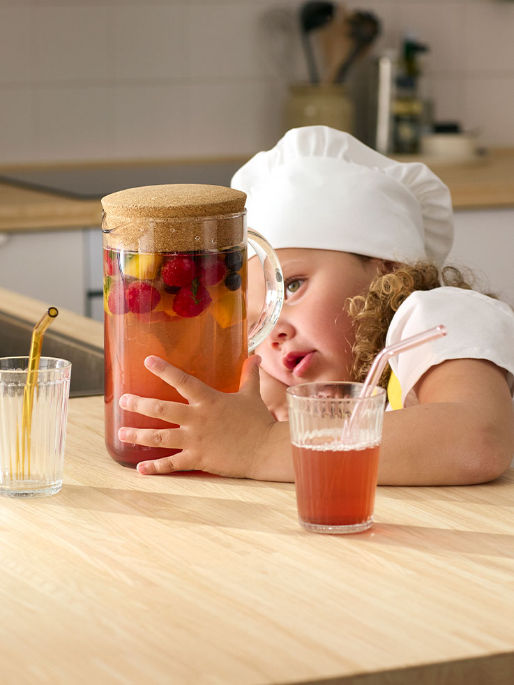 A little girl dressed in an apron and a chef’s hat, longingly looking at a jug of water and lots of berries and fruit.
