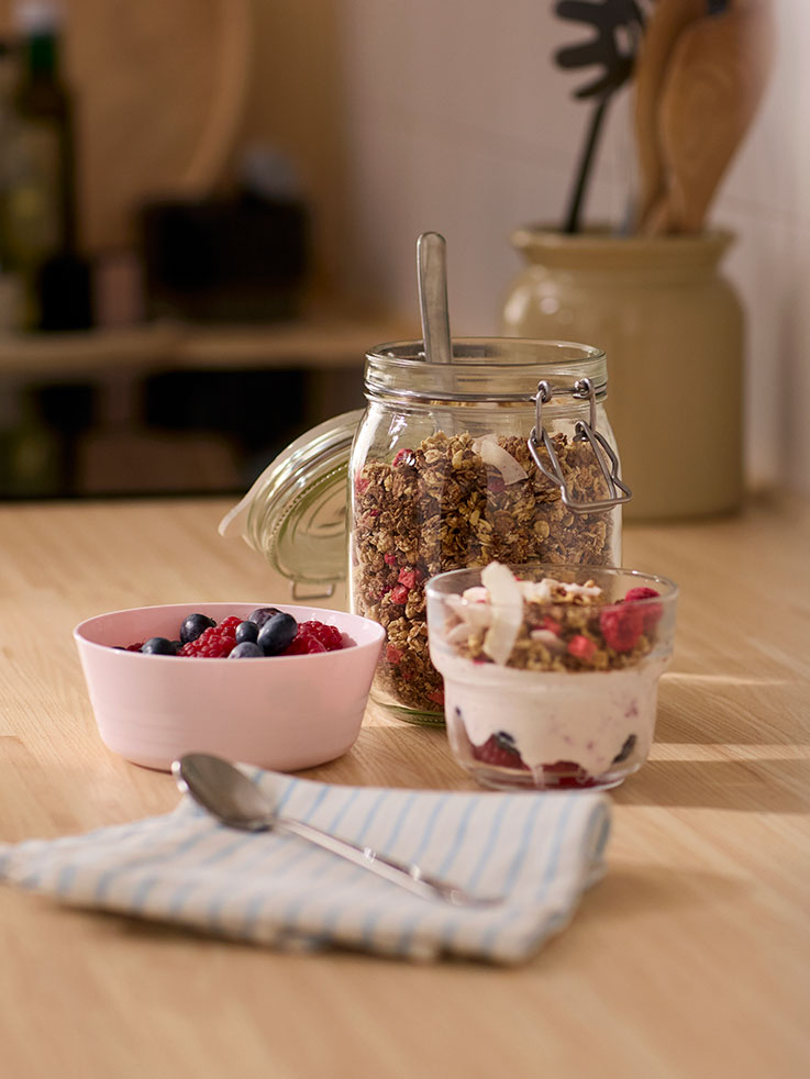 A little girl dressed in an apron and a chef’s hat, longingly looking at a jug of water and lots of berries and fruit.