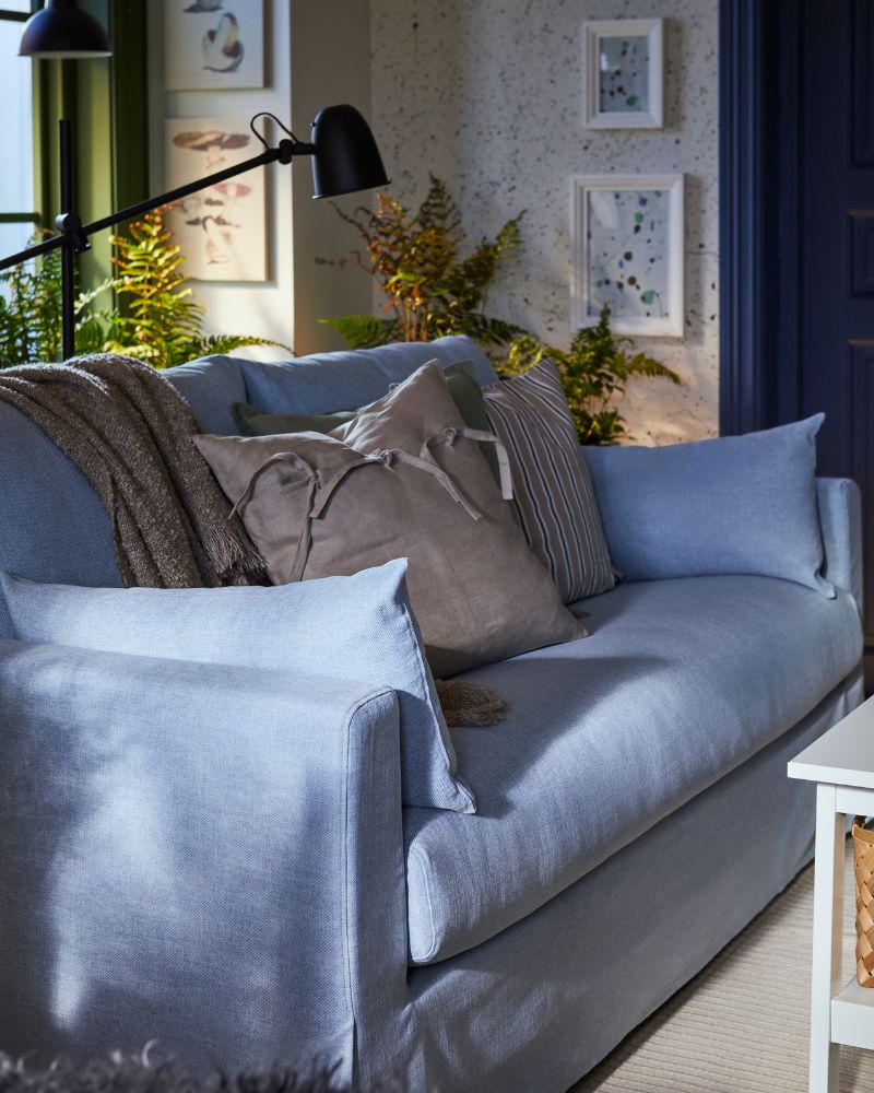 A pale blue HYLTARP 2-seat sofa with a throw and two extra cushions on it. One cushion has a beige AINA cushion cover.