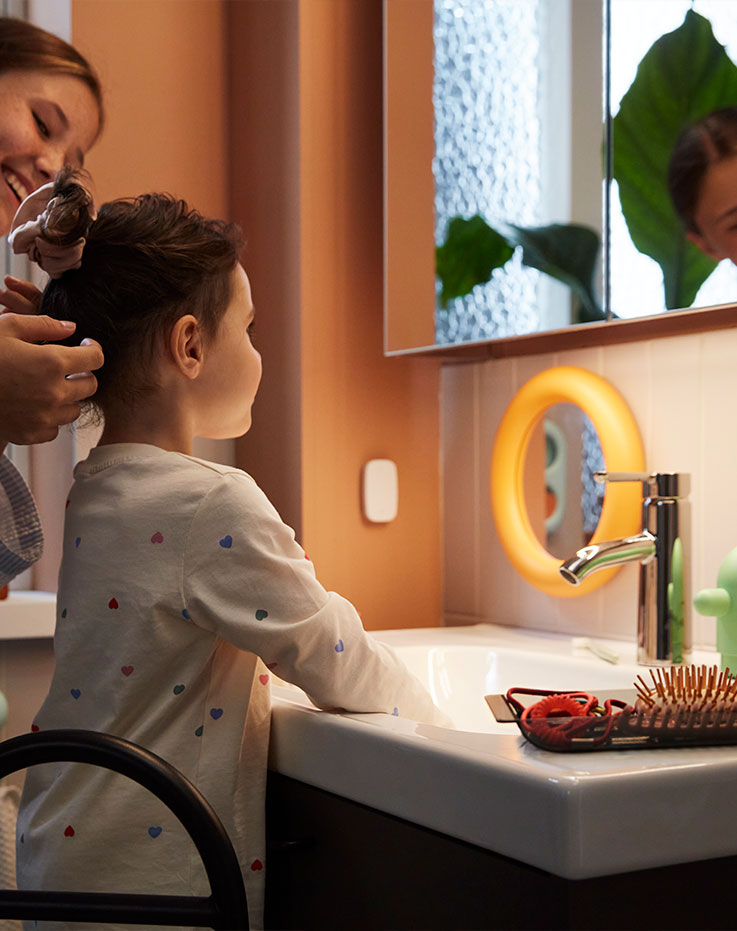A bathroom with a LETTAN mirror cabinet showing a  child getting ready with an adult by a sink with a green  plant in the back.