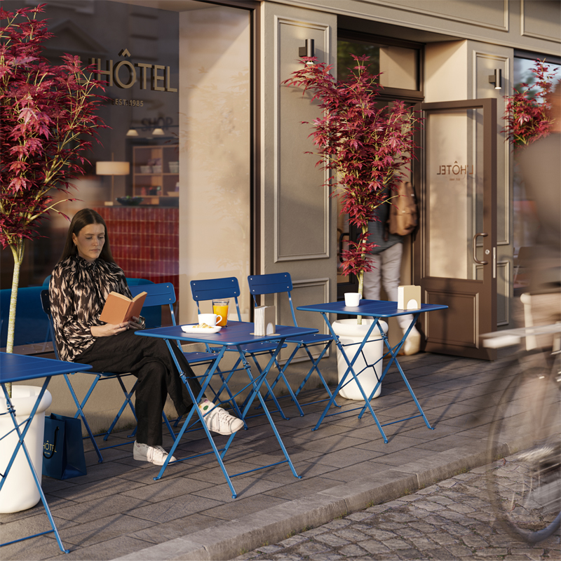 Outdoor area outside a hotel lobby with dark blue SUNDSÖ tables and chairs with a person reading whilst enjoying coffee.