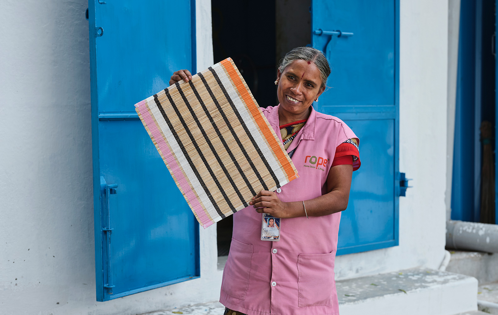 A person in pink clothes stands smiling in front of a pair of blue doors and holds up a striped MÄVINN place mat.