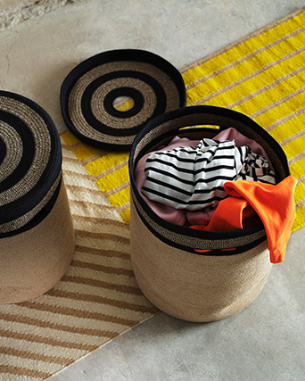 Two MÄVINN laundry baskets stand on a yellow rug. One has a lid on, the other is filled with laundry with the lid next to it.