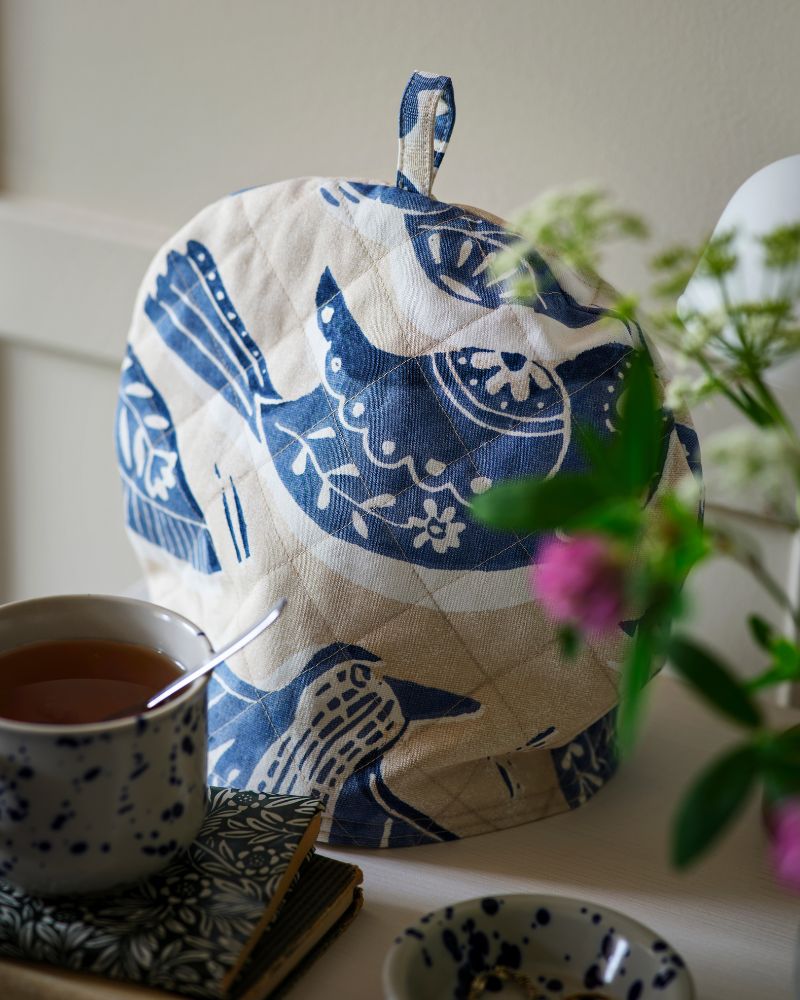 A tea cosy crafted from beige and blue BITTERSÖTA pre-cut fabric sits atop a teapot, next to a mug and some notebooks.