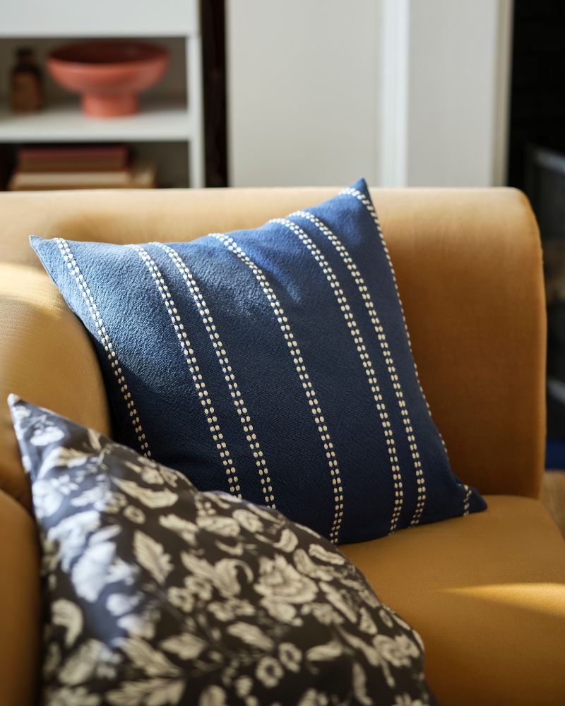 A striped KRUTBRÄNNARE cushion cover in blue off- white sits on a beige STOCKHOLM two-seat sofa next to a patterned cushion.