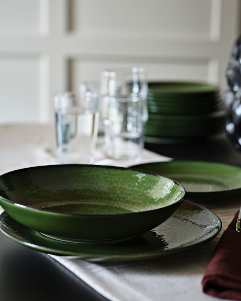 A green PELARKAKTUS deep plate sits on a table beside glassware and stacked plates in a stylish dining setting.