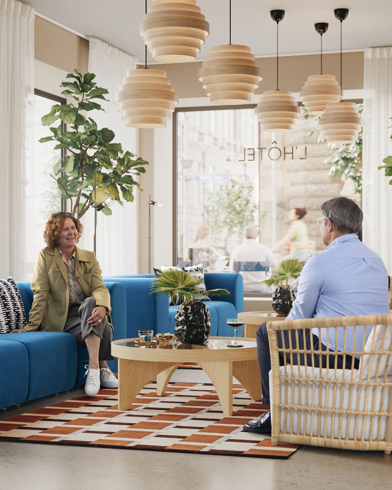 Hotel lobby scene with a grey/white STOCKHOLM 2025 rattan armchair, a sofa, coffee table, and two people sitting and talking.