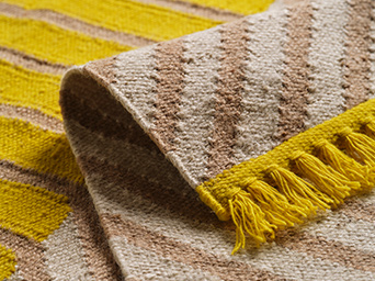 A close-up of the yellow fringes of a flatwoven jute/wool MÄVINN rug with a yellow and beige striped pattern.