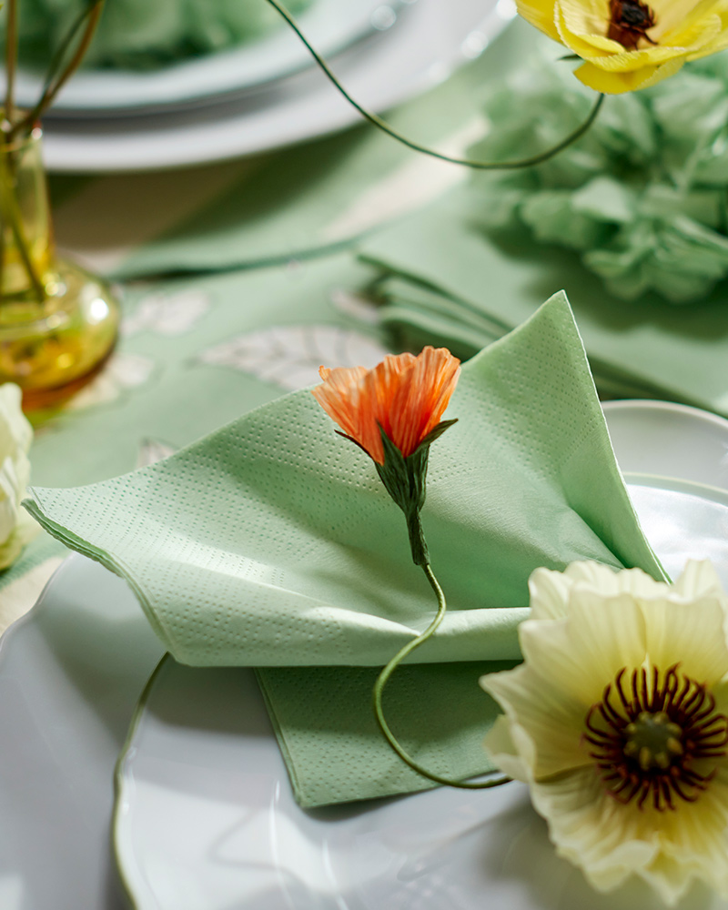 A green FANTASTISK paper napkin, on top of UPPLAGA side plate and plate, decorated with a SMYCKA artificial spray magnolia.