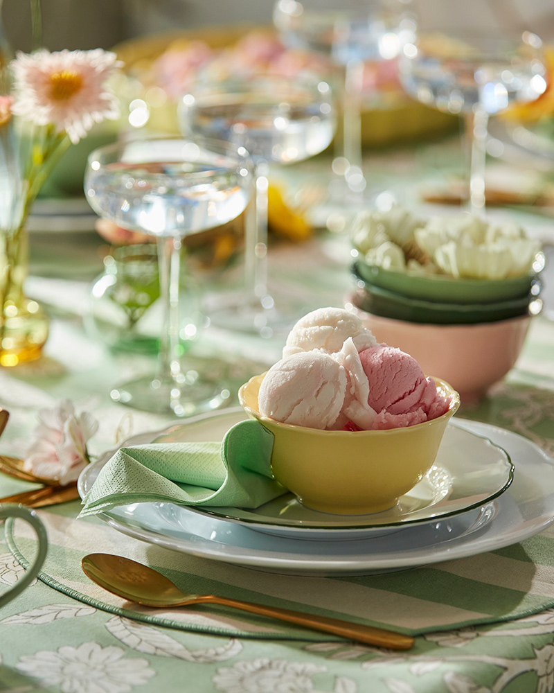 A yellow UPPLAGA bowl holds pink and white ice cream, surrounded by colourful table settings of flowers and glassware.