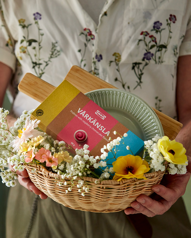 A person holds a SMÖRFISK breadbasket, filled with a VÅRKÄNSLA praline gift box, decorated with SMYCKA artificial sprays.