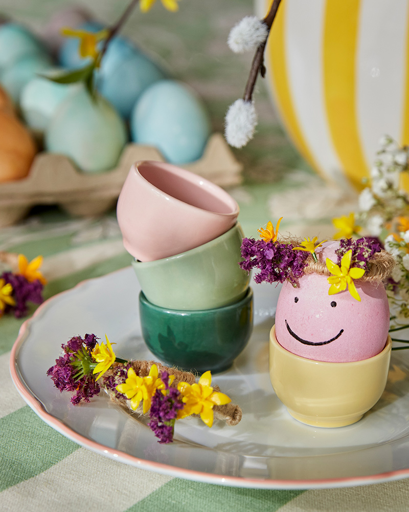 An Easter egg painted like a smiley face, in a pastelcoloured IKEA 365 egg cup on top of an UPPLAGA plate.