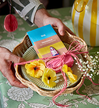 A person holds a SMÖRFISK breadbasket, with a VÅRKÄNSLA bag of chocolate bunnies, decorated with a SMYCKA artificial spray.