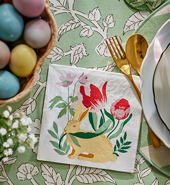 A close-up of an Easter bunny-patterned TJÄRLEK paper napkin, next to an UPPLAGA plate on a green SMÖRFISK tablecloth.