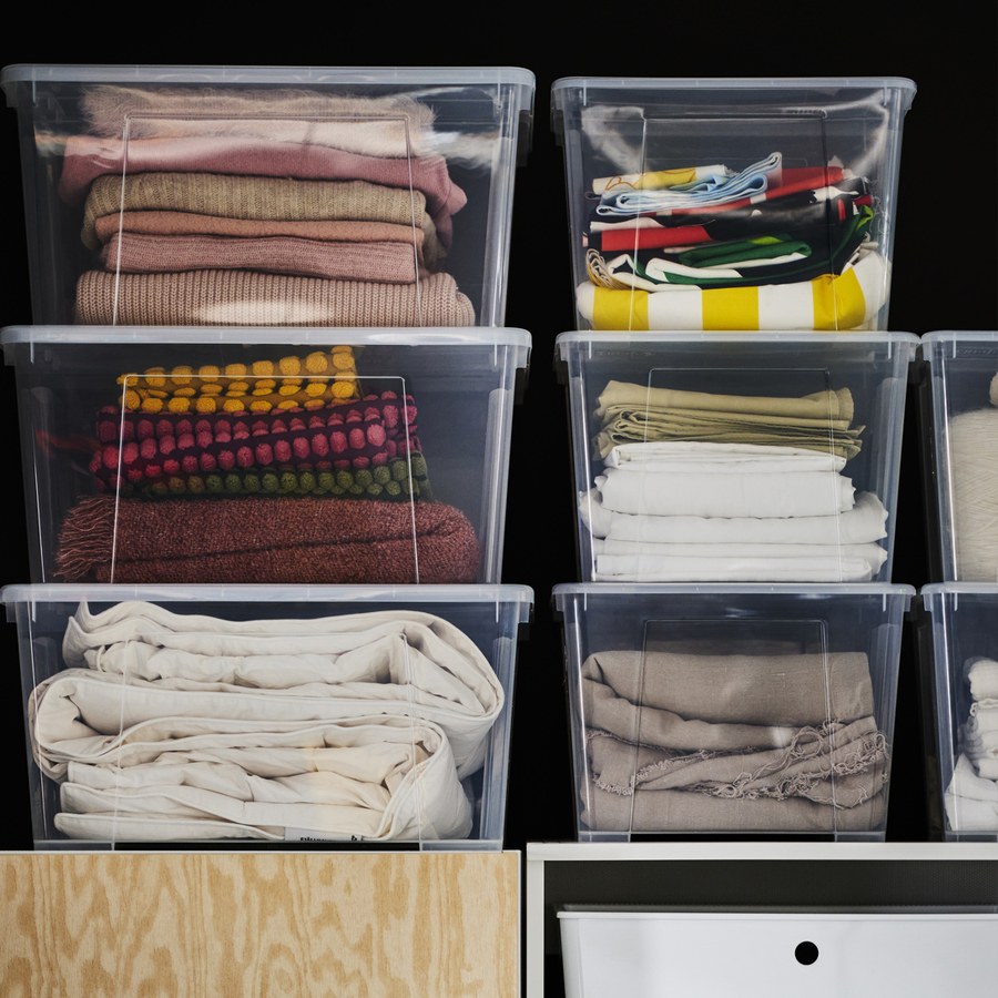 Stacked clear storage boxes containing neatly folded clothes and textiles, placed on shelving units.