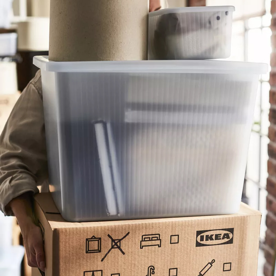 Person carrying a cardboard box topped with transparent storage boxes during a move.