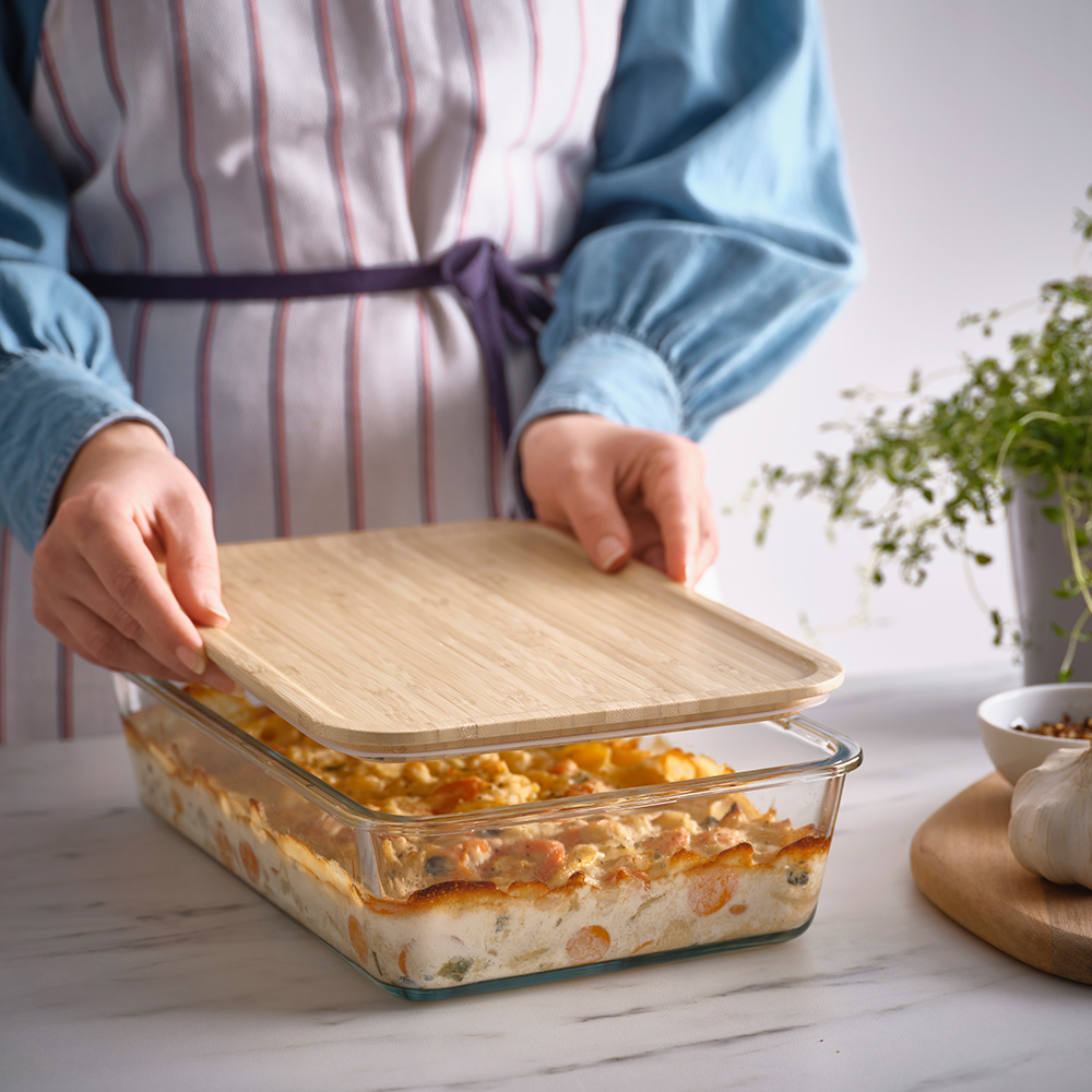 A person in a striped apron places a bamboo lid on an IKEA 365+ glass food container filled with a casserole.