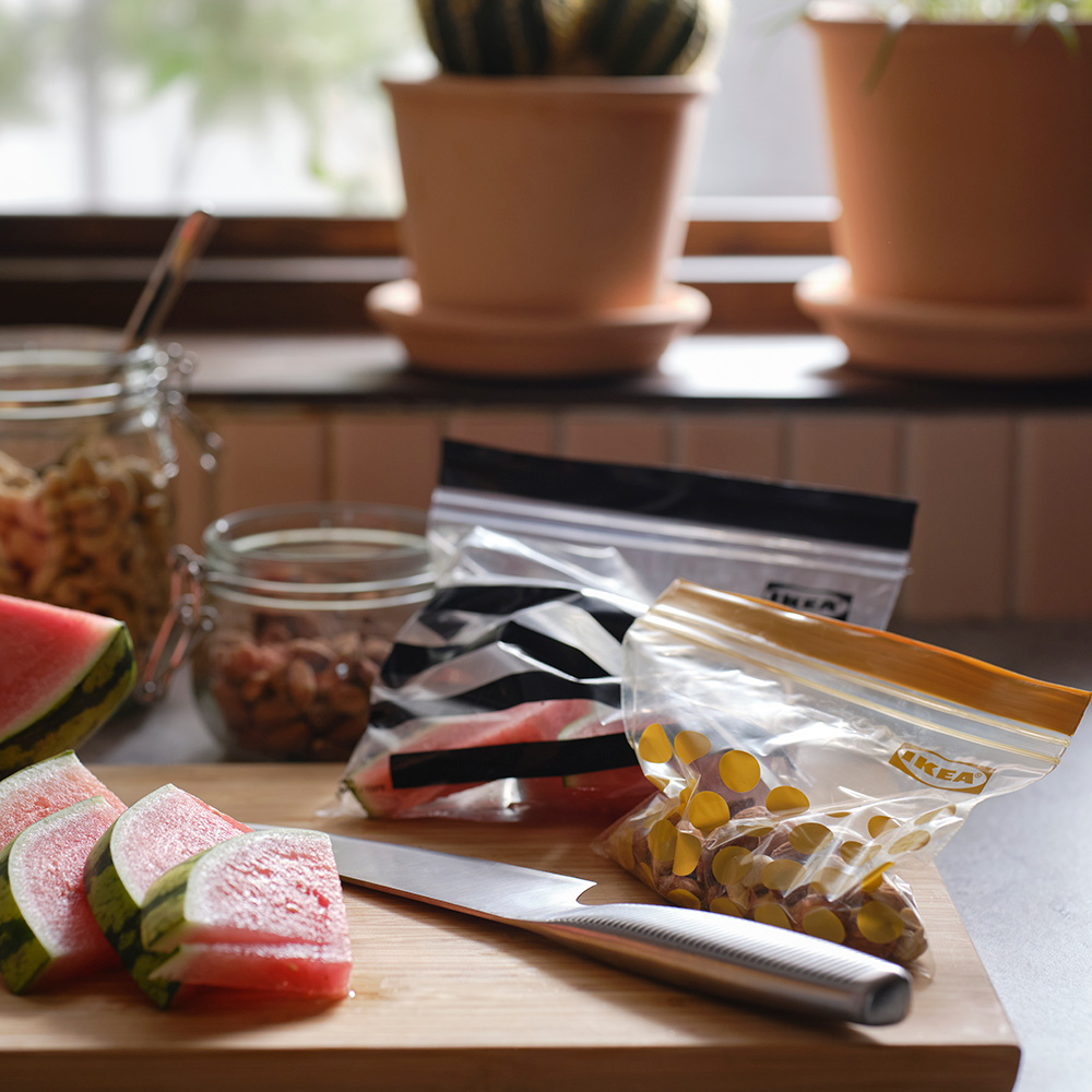 Slices of watermelon, a knife and two different sized ISTAD plastic resealable bags are on a cutting board in a kitchen.
