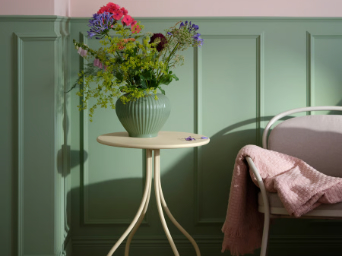 Una mesa auxiliar TÅNEBRO de color amarillo pálido sostiene un jarrón con flores junto a un sillón en una habitación con paredes verdes y luz tenue.
