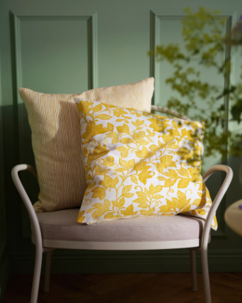 Una funda de cojín FJUNKLOCKA amarilla y blanca descansa sobre un sillón claro, rodeada de paredes verdes y una luz suave.