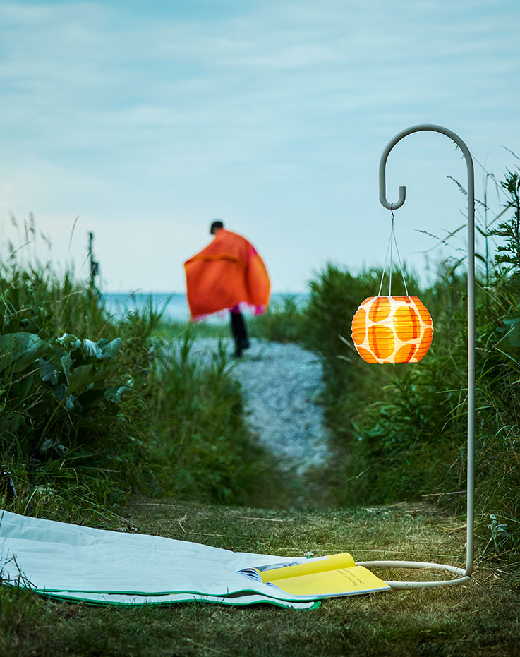 A garden evening setting features a beige SOLVINDEN lamp stand with an illuminated orange lampshade and a person walking.