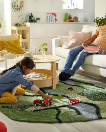 En una sala de estar iluminada por el sol, un niño se sienta en una alfombra GREJSIMOJS y juega con un tren de juguete mientras su padre se sienta en el sofá.