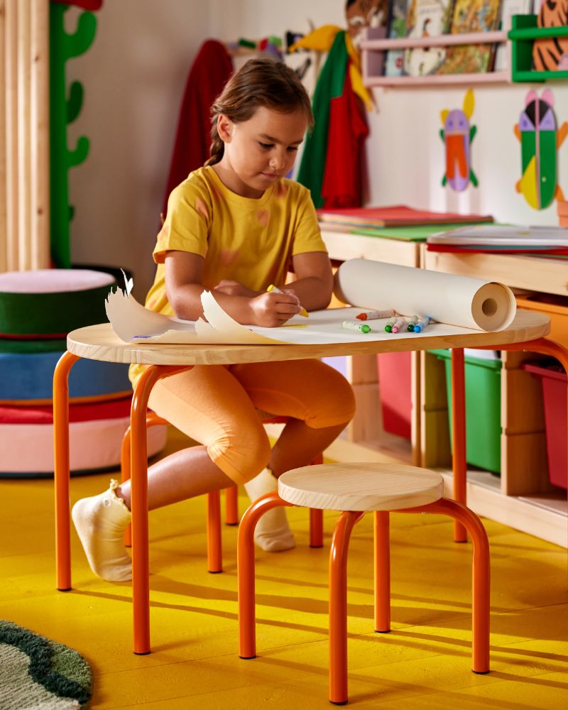 A child works with paper and art supplies while using a GRESIMOJS children’s table and stool in a bright and colourful room.