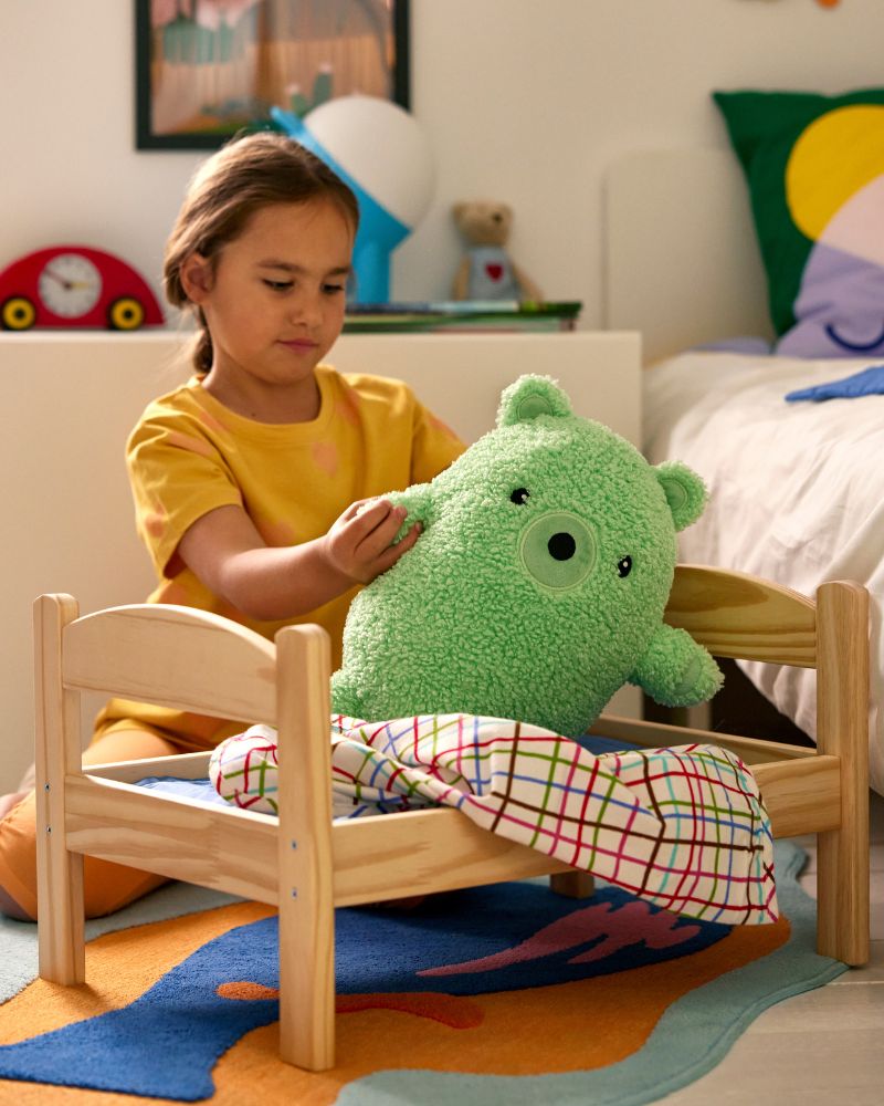 A child plays with a green GREJSIMOJS soft toy bear and a toy bed with blanket in a colourful and cosy kid’s Open in Komponera  room.