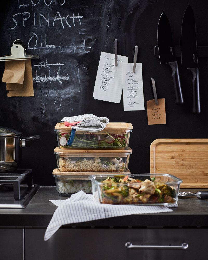 IKEA 365+ glass food containers with bamboo lids are on a dark kitchen worktop. A black chalkboard is behind the containers.