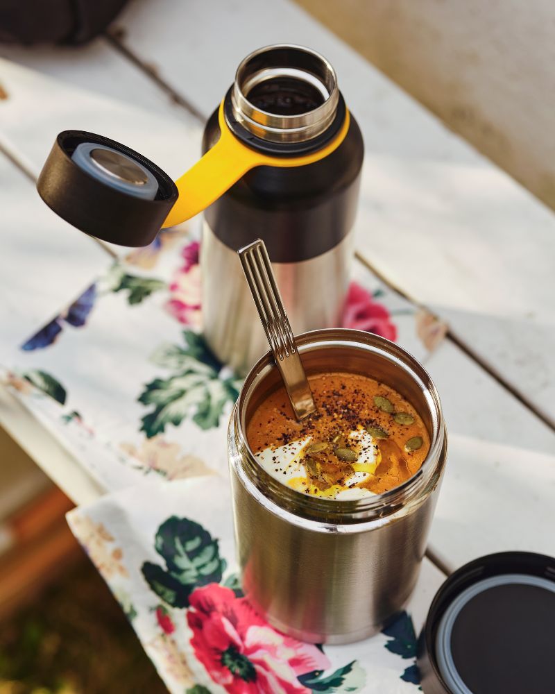An open EFTERFRÅGAD food vacuum flask with soup and a spoon in it and its lid are on top of a floral napkin on a tabletop.