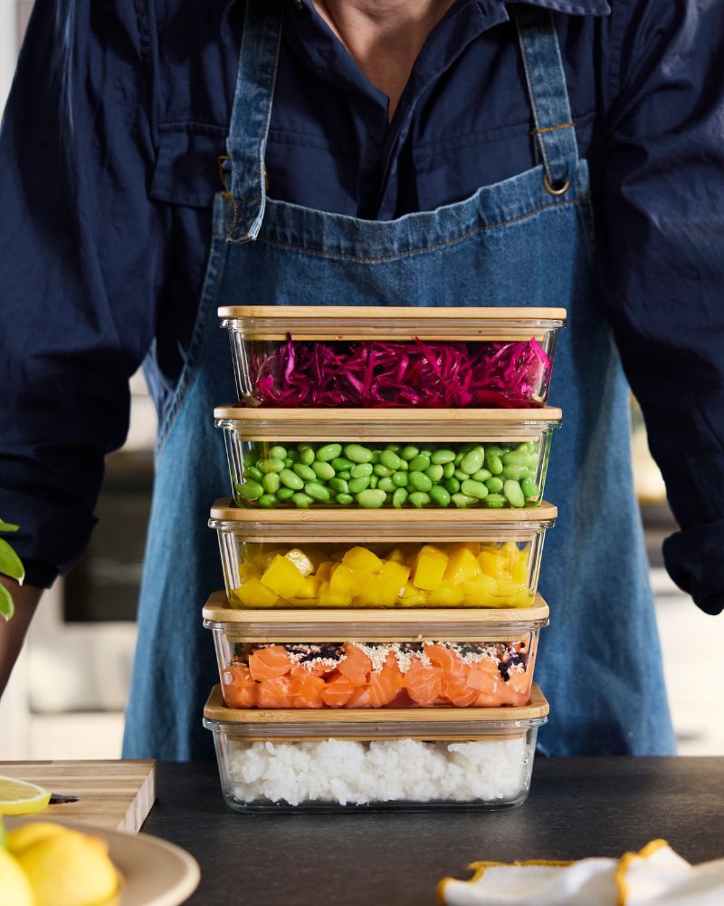 A person wearing a denim apron stands behind a stack of IKEA 365+ glass food storage containers filled with prepared food.