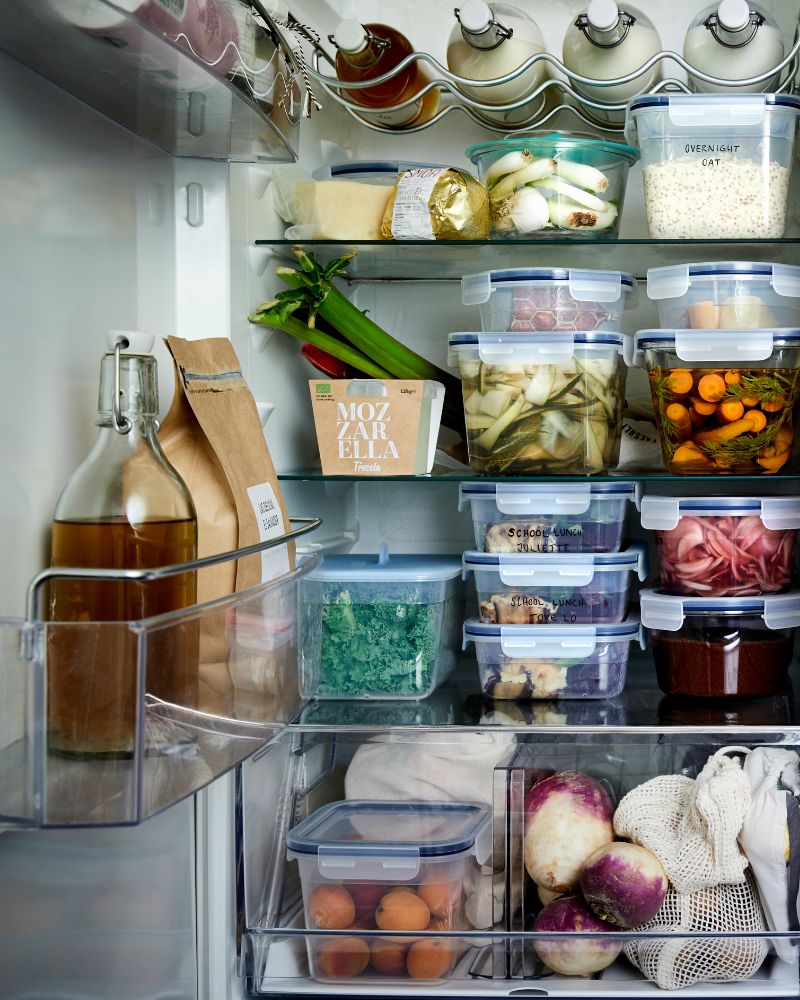 An open refrigerator is filled with glass bottles and IKEA 365+ plastic food storage containers full of food.