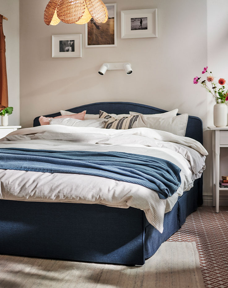 A blue grey HUMLEMOTT throw lies across a RAMNEFJÄLL upholstered bed that’s covered in SCHERSMIN and SOLFIBBLA bed linen.