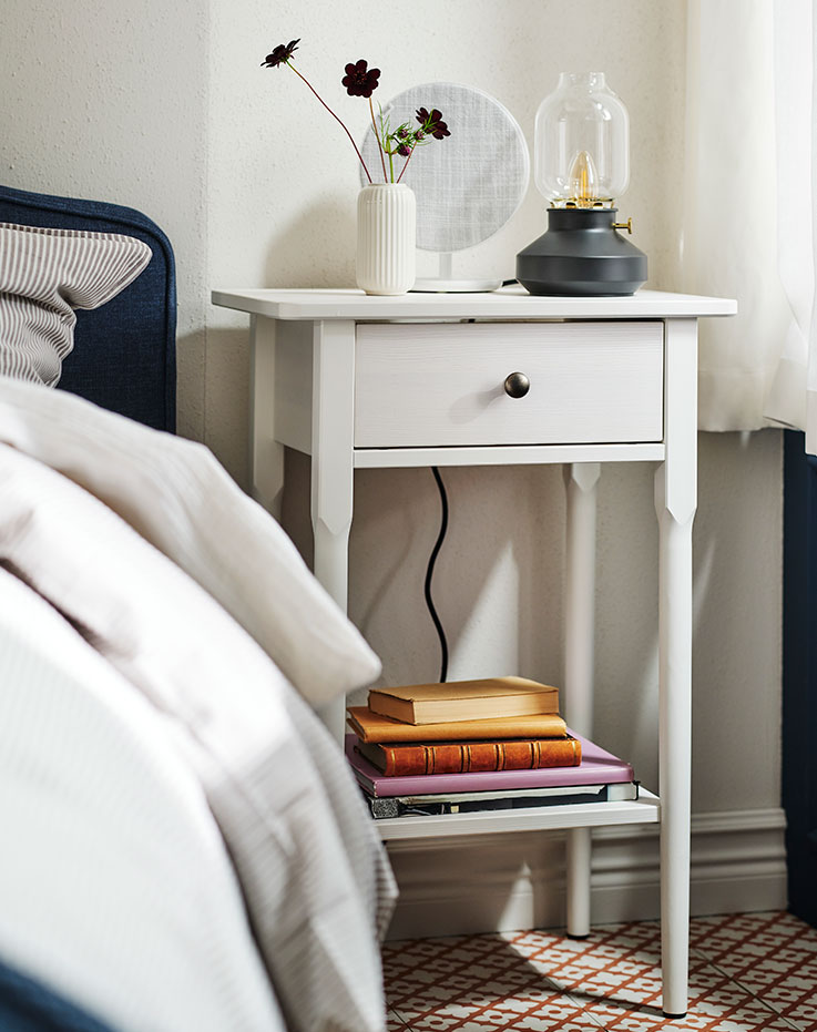 A BLÄSTÅSEN bedside table by a RAMNEFJÄLL bed with a SOLSKYDD Bluetooth speaker on top and some books on the lower shelf.