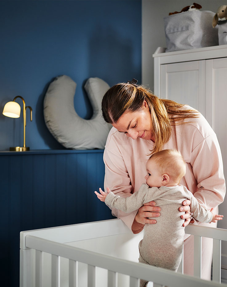 A person picks up a baby from a white SUNDVIK cot in the corner of a dark blue bedroom with a white wardrobe behind them.