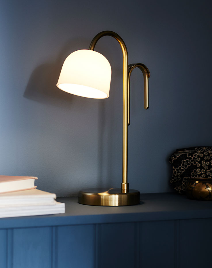 A brass/white glass TVÅMASTAD LED portable lamp that is switched on placed on a dark blue ledge with some books beside it.