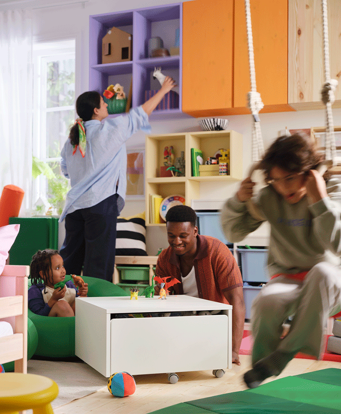 Family playing in a colorful room with a white storage table, purple wall cubbies, and an orange cabinet while a child swings in the foreground.