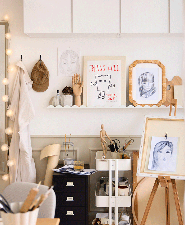 Art studio corner with a white rolling cart, an easel with a portrait, and a wall shelf displaying sketches, a wooden hand, and a brown cap.
