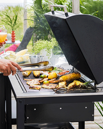 Primer plano de una barbacoa de carbón negra con mazorcas de maíz, piña y comida en papel de aluminio cocinándose sobre la parrilla abierta.