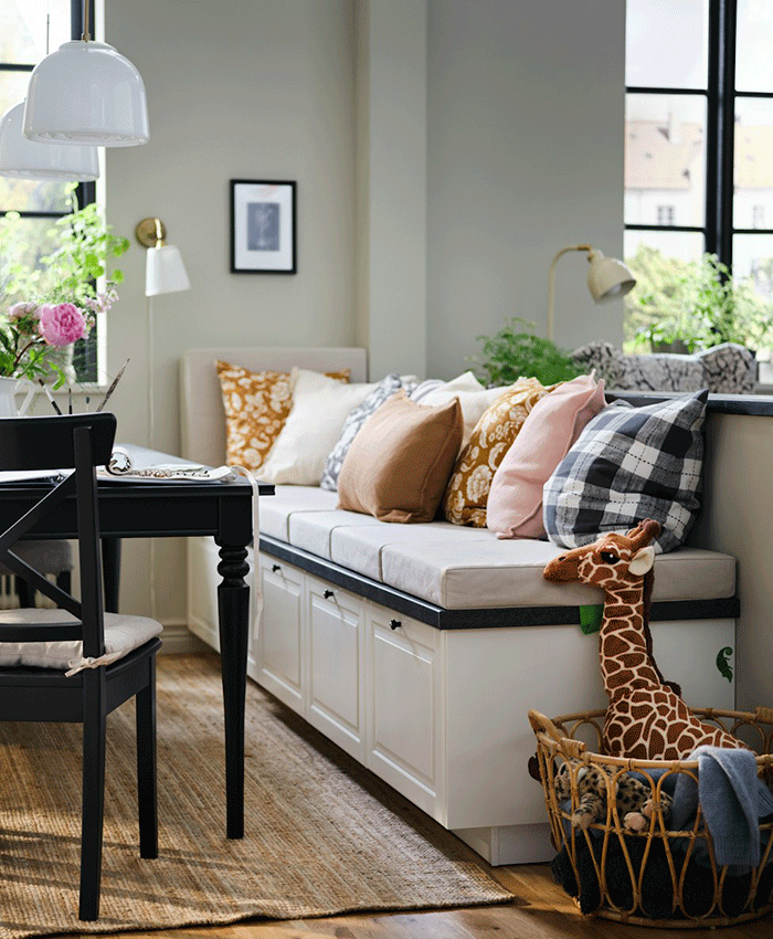 White storage bench with cushions in earthy tones and patterns, used as seating for a black dining table. A giraffe plush toy sits in a wicker basket nearby.