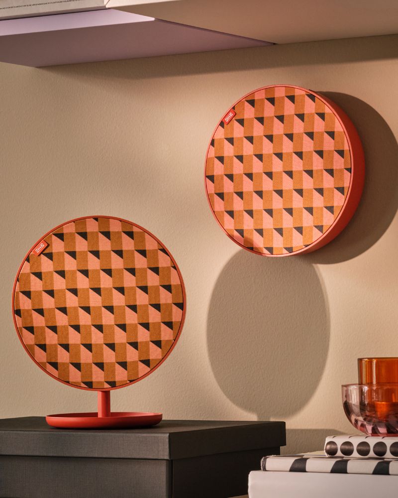 A living-room corner with two orange SOLSKYDD portable Bluetooth speakers, one mounted on a stand, the other on the wall.