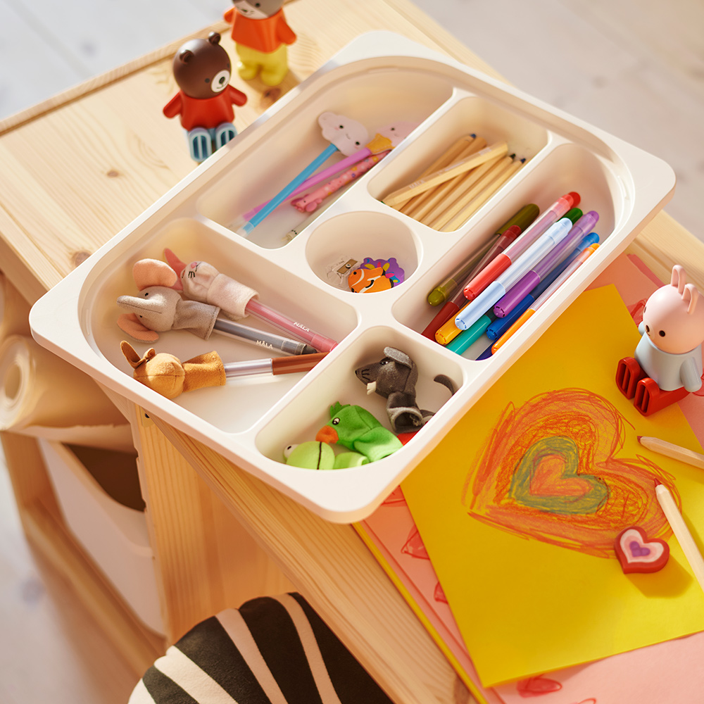 Una bandeja TROFAST blanca con compartimentos que contiene bolígrafos y lápices está sobre una encimera TROFAST, con un dibujo al lado.