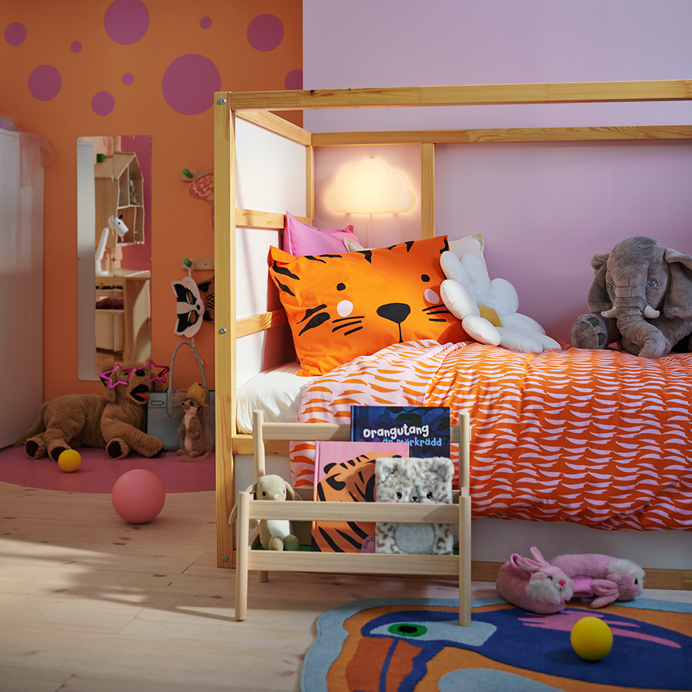 Una cama reversible KURA en un colorido dormitorio infantil, con peluches encima y un expositor de libros FLISAT al frente.