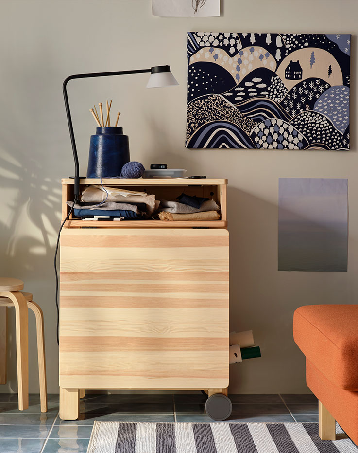 A sunlit living room with a pine RESARÖ mobile drop leaf table in folded mode, by a wall and next to stacked KYRRE stools.
