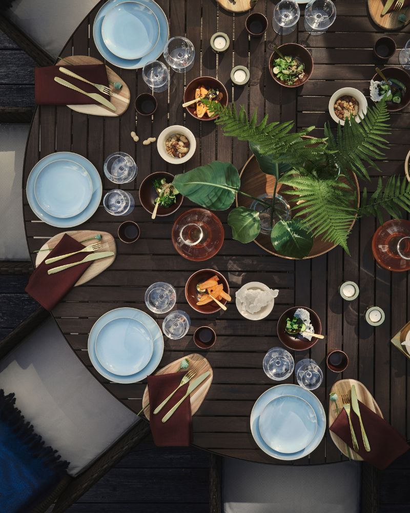 A dining table set outdoors with dark grey VITTSKÄR round table arranged with light blue FÄRGKLAR dinnerware and plants.