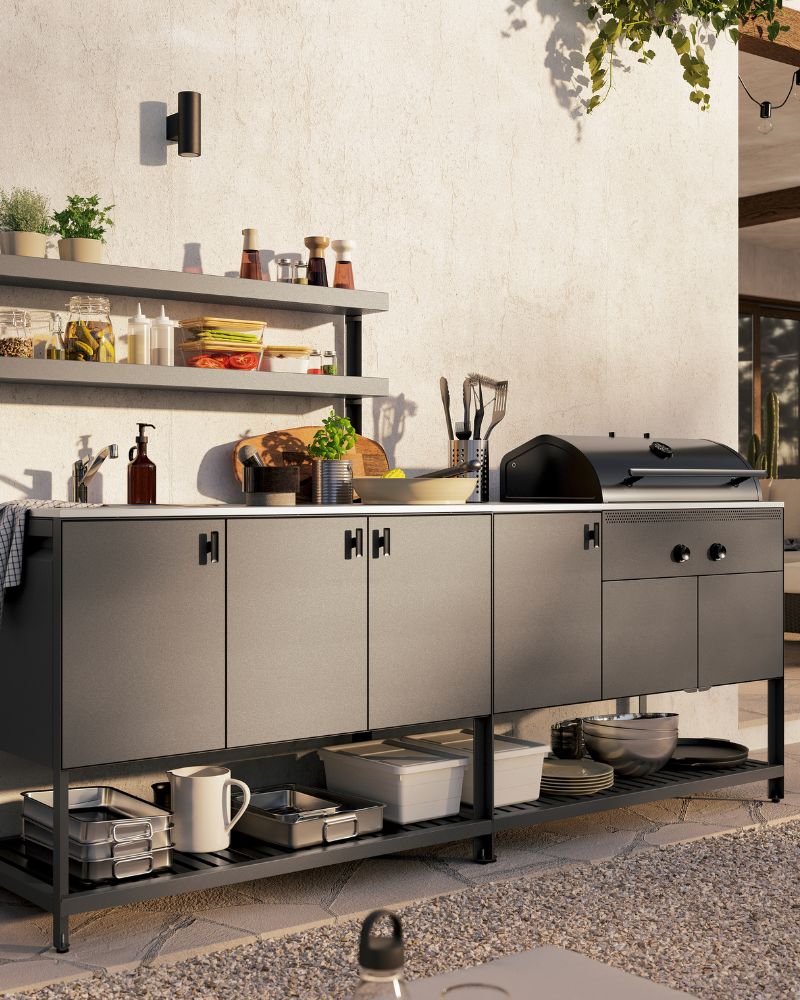 A dark grey BÅTSKÄR outdoor kitchen with charcoal barbecue. Above the closed cabinets are two open shelves with fresh herbs.