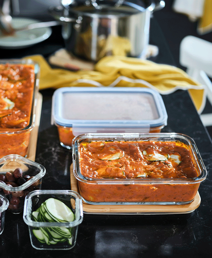 Glass IKEA 365+ food containers filled with vegetable stew and sliced zucchini on a dark kitchen counter with a yellow cloth.