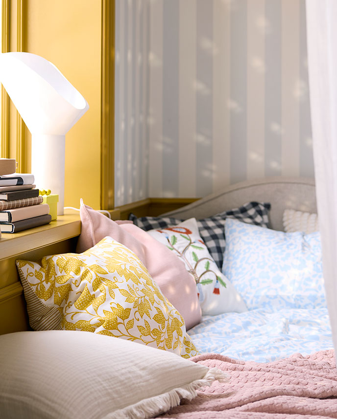 A close-up of the FJUNKLOCKA cushion cover in yellow and white, placed among various textured pillows on a cosy bed.