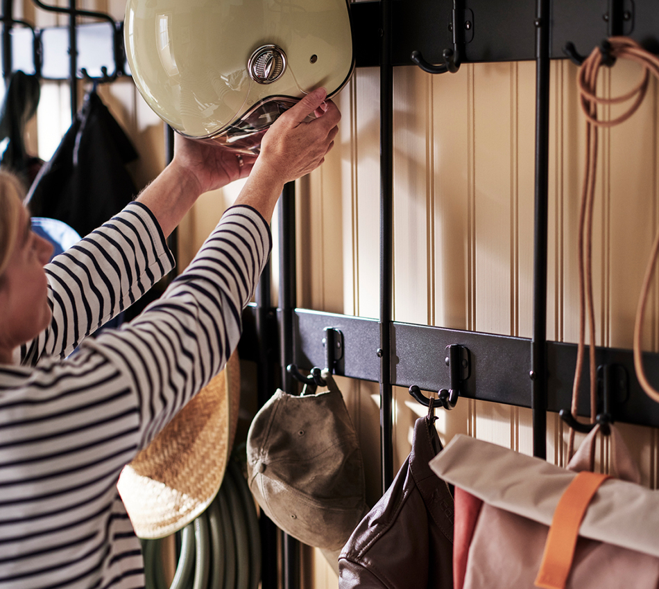 Close-up of hands placing a cream-colored motorcycle helmet on a black metal wall rack with hooks holding a cap and a brown leather jacket.