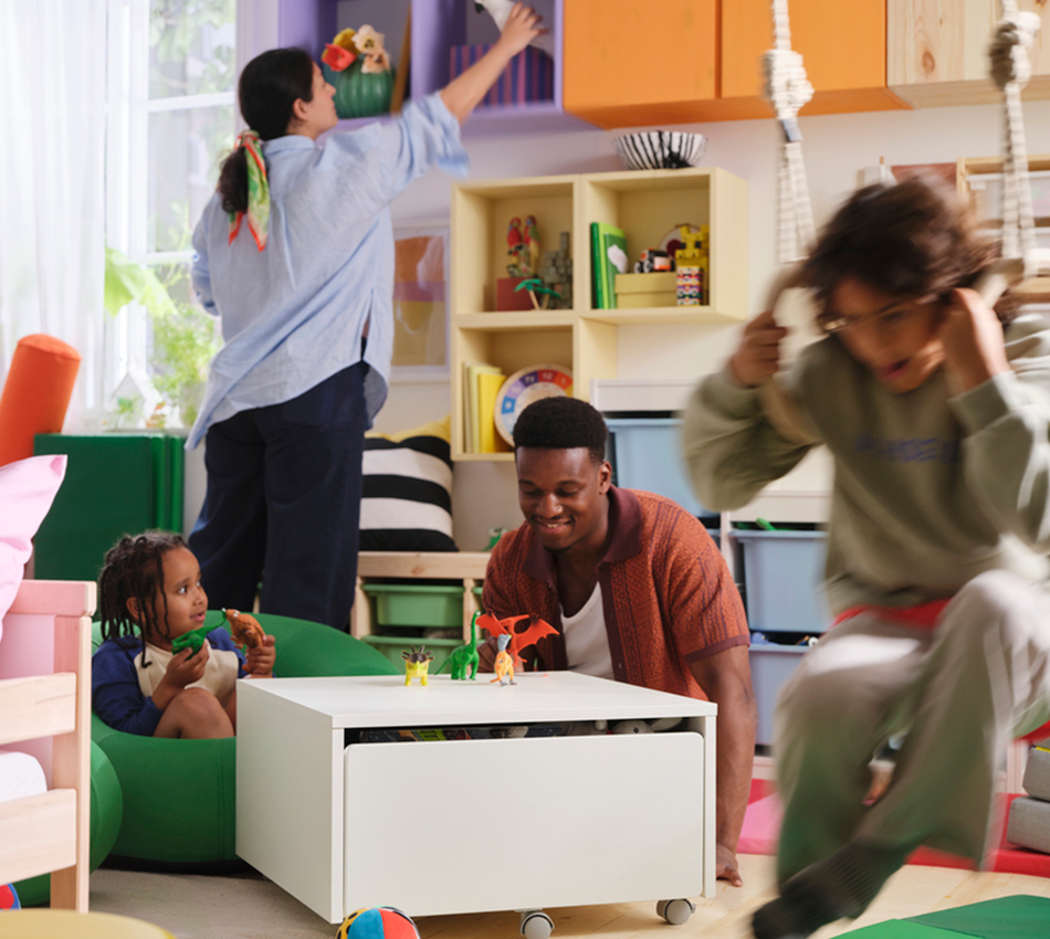 Family playing in a colorful room with a white storage table, purple wall cubbies, and an orange cabinet while a child swings in the foreground.